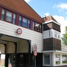 Poortgebouw met twee winkels met bovenwoningen, met veel hout zoals de zware kolommen in poort, bouwstijl Amsterdamse School, ligging aan Purmerweg centraal deel van Tuindorp Nieuwendam