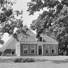 Boerderij met groot schuurdak en dwars voorhuis met omlijste ingang en geblokte kroonlijst onder schilddak met tweedelige dakkapel en twee hoekschoorstenen met versierde borden