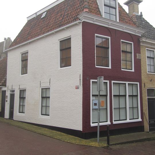 Eenvoudig hoekpand met gebroken dak en gevelsteen in de verdieping, waar boven de vensters bogen bewaard zijn