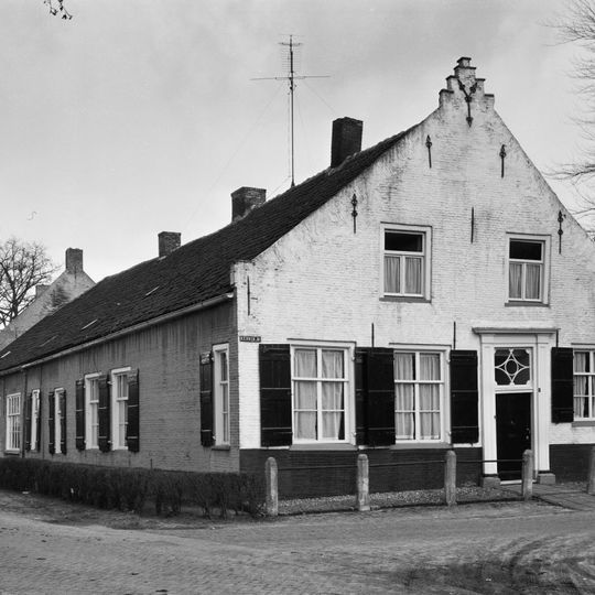 Huis onder met pannen belegd zadeldak tussen topgevels waarvan de korte voorgevel met een getrapte top; zesdelige schuiframen; ingang met pilasters en kroonlijst. Lelie-ankers
