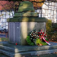 Quaderförmiger Steinblock (auf zweistufigem Podest stehend). Seitlich Tafeln mit den Namen von Gefallenen oder Vermissten. Darauf ein überlebensgroßer steinerner Soldatenhelm auf einem Kranz.