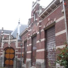 Schuin in de rooilijn staand pand, waarin gehuisvest de Herenkamer, onder afgeplat schilddak met leien gedekt, opgetrokken in baksteen, afgewisseld met natuurstenen blokken en banden.