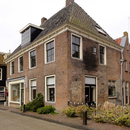 Aan drie zijden vrijstaand breed pand met jongere pui en smalle geprofileerde rondgaande daklijst met blokjes onder fors schilddak met jongere dakkapel