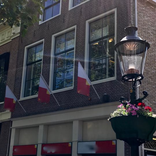 Hoog pand tegen klokgevel met natuurstenen aanzetkrullen en kroonlijst met emblemen onder zadeldak
